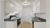This single-floor unit offers a clean, functional layout with a consistent neutral color palette throughout. The galley-style kitchen is efficiently designed with white shaker cabinetry lining both walls, dark speckled countertops, and a full suite of appliances including an electric range, refrigerator, and dishwasher. A stainless steel sink and under-cabinet range hood add practical utility, while wood-look plank flooring ties the space together with a warm tone.

The living area is generously proportioned, featuring plush wall-to-wall carpeting, a baseboard heating unit, and a large sliding door dressed with vertical blinds that allows natural light to fill the room. A ceiling fan with a light fixture is present in a secondary area near the entryway, which also benefits from wood-look flooring and built-in closet storage.

The bedroom is carpeted and offers a roomy feel, complemented by a double-door closet providing ample storage. The full bathroom maintains the space's clean aesthetic with an all-white finish, a tub-shower combination with tiled surround, a single vanity with a large mirror, and wood-look flooring. Overall, the unit presents a bright, move-in-ready atmosphere with practical finishes throughout.