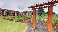 The community gardens offer a well-organized outdoor growing space designed to foster resident engagement with nature and sustainable gardening. At the heart of the garden, a striking deep-red wooden pergola serves as a welcoming entryway, featuring lattice side panels and an open overhead framework that frames views of the garden beds beyond. Mulched pathways wind between the numerous raised planting beds, which are constructed from rich, warm-toned timber and arranged in a thoughtful grid-like layout that maximizes growing space while maintaining ease of access.

The beds themselves support a diverse array of plantings, including tomatoes, squash, sunflowers, leafy greens, and various other vegetables and flowering plants, reflecting active, productive use throughout the growing season. The open-air setting provides full sun exposure, essential for healthy plant growth, while the surrounding landscape of manicured lawn and mature trees lends a pleasant, park-like atmosphere. The Tudor-style multi-story residential buildings visible in the background provide an architectural frame to the garden, reinforcing its role as a shared community amenity. Overall, the space conveys a welcoming, industrious character ideal for residents seeking a hands-on connection to gardening.