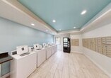This well-maintained laundromat offers a clean and functional environment designed to accommodate a variety of laundry needs. The space is defined by its distinctive tray ceiling painted in a calming aqua-blue tone, complemented by recessed lighting that keeps the interior bright and welcoming. The walls feature a matching light blue accent color, while crisp white trim and light-toned porcelain tile flooring tie the design together for a fresh, modern appearance.

A row of top-loading washing machines lines one wall, providing ample washing capacity. Opposite, a bank of stacked dryers offers efficient drying options. A built-in folding counter with a dark laminate surface provides a dedicated space for sorting and folding laundry. A wall-mounted air conditioning unit ensures year-round comfort for users.

A vending machine stocked with snacks and beverages offers added convenience during longer visits. Along one wall, an extensive array of individual mailboxes serves residents, making this space a practical multi-use facility. A window near the washing machines allows natural light to filter in, adding to the airy atmosphere. Security cameras are present throughout, ensuring a safe environment. The space is accessible via a glass-panel entry door, completing this efficient and thoughtfully designed community laundry facility.