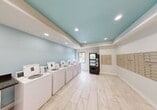 This well-maintained laundromat offers a clean and functional environment designed to accommodate a variety of laundry needs. The space is defined by its distinctive tray ceiling painted in a calming aqua-blue tone, complemented by recessed lighting that keeps the interior bright and welcoming. The walls feature a matching light blue accent color, while crisp white trim and light-toned porcelain tile flooring tie the design together for a fresh, modern appearance.

A row of top-loading washing machines lines one wall, providing ample washing capacity. Opposite, a bank of stacked dryers offers efficient drying options. A built-in folding counter with a dark laminate surface provides a dedicated space for sorting and folding laundry. A wall-mounted air conditioning unit ensures year-round comfort for users.

A vending machine stocked with snacks and beverages offers added convenience during longer visits. Along one wall, an extensive array of individual mailboxes serves residents, making this space a practical multi-use facility. A window near the washing machines allows natural light to filter in, adding to the airy atmosphere. Security cameras are present throughout, ensuring a safe environment. The space is accessible via a glass-panel entry door, completing this efficient and thoughtfully designed community laundry facility.