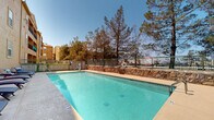 Located in Las Cruces, New Mexico, this outdoor amenity area centers around a generously sized swimming pool with clear, turquoise water. The pool is equipped with safety features including a life ring station and entry handrails, ensuring a secure environment for all users. Along one side of the pool deck, a row of blue cushioned chaise lounges provides ample space for sunbathing and relaxation, with additional seating and a shaded umbrella area nearby. A kettle-style grill adds an outdoor entertaining element to the patio space. The area is enclosed by a combination of black iron fencing and a rustic stacked-stone retaining wall, complemented by mature pine trees that offer both privacy and natural shade. The warm, adobe-toned building facade rises adjacent to the pool deck, featuring multiple balconies and windows that overlook the outdoor space. The wide concrete surround provides generous room for movement around the pool, while the open sky above allows for abundant natural sunlight throughout the day. The overall atmosphere is well-suited for outdoor leisure, capturing the sunny, relaxed character of the New Mexico desert climate.