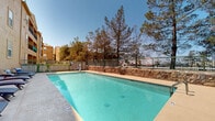 Located in Las Cruces, New Mexico, this outdoor amenity area centers around a generously sized swimming pool with clear, turquoise water. The pool is equipped with safety features including a life ring station and entry handrails, ensuring a secure environment for all users. Along one side of the pool deck, a row of blue cushioned chaise lounges provides ample space for sunbathing and relaxation, with additional seating and a shaded umbrella area nearby. A kettle-style grill adds an outdoor entertaining element to the patio space. The area is enclosed by a combination of black iron fencing and a rustic stacked-stone retaining wall, complemented by mature pine trees that offer both privacy and natural shade. The warm, adobe-toned building facade rises adjacent to the pool deck, featuring multiple balconies and windows that overlook the outdoor space. The wide concrete surround provides generous room for movement around the pool, while the open sky above allows for abundant natural sunlight throughout the day. The overall atmosphere is well-suited for outdoor leisure, capturing the sunny, relaxed character of the New Mexico desert climate.