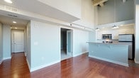 This two-story space showcases an open, airy design highlighted by dramatically high ceilings that emphasize the generous vertical scale of the interior. The main living area features rich, warm-toned hardwood flooring that runs throughout, lending a polished yet inviting character to the space. Soft, neutral wall tones in a muted blue-gray palette create a calm and cohesive atmosphere across the floor.

A sliding frosted-glass door provides a stylish and functional partition, leading to an adjacent carpeted room that offers a quieter, more enclosed setting. Exposed structural beams are visible at the upper level, adding an industrial architectural detail that contrasts tastefully with the otherwise clean, modern finishes.

The kitchen is partially open to the main living area and features white cabinetry, stainless steel appliances including a built-in microwave, and a peninsula counter with bar seating overhang — ideal for casual dining or entertaining. A pendant light fixture hangs above, providing focused illumination. The upper level overlooks the main floor, reinforcing the spacious, loft-like quality of the overall design.