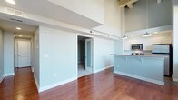 This two-story space showcases an open, airy design highlighted by dramatically high ceilings that emphasize the generous vertical scale of the interior. The main living area features rich, warm-toned hardwood flooring that runs throughout, lending a polished yet inviting character to the space. Soft, neutral wall tones in a muted blue-gray palette create a calm and cohesive atmosphere across the floor.

A sliding frosted-glass door provides a stylish and functional partition, leading to an adjacent carpeted room that offers a quieter, more enclosed setting. Exposed structural beams are visible at the upper level, adding an industrial architectural detail that contrasts tastefully with the otherwise clean, modern finishes.

The kitchen is partially open to the main living area and features white cabinetry, stainless steel appliances including a built-in microwave, and a peninsula counter with bar seating overhang — ideal for casual dining or entertaining. A pendant light fixture hangs above, providing focused illumination. The upper level overlooks the main floor, reinforcing the spacious, loft-like quality of the overall design.
