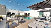 This contemporary rooftop amenity deck is a standout outdoor retreat defined by clean architectural lines, bright white parapet walls, and an open-air design that maximizes exposure to natural light and sky. At the heart of the space is a pristine swimming pool with vivid turquoise water, framed by wide-format tile decking that creates a sleek, resort-inspired aesthetic. Multiple stainless steel entry rails provide easy pool access, and raised planter beds filled with drought-tolerant greenery add a natural contrast to the modern hardscape. A generous sunbathing zone surrounds the pool, outfitted with cushioned chaise lounges positioned beneath large shade umbrellas, offering both sun and shelter options throughout the day. A dedicated lounge area along the rooftop perimeter features a long, neutral-toned sectional sofa paired with round terrazzo side tables, all situated directly against glass railings that frame sweeping views of the city and distant mountains. Overhead, globe string lights are strung across the deck, lending a warm, inviting ambiance for evening gatherings. A partially covered cabana area provides additional seating and shade, while the elevated position of the space delivers unobstructed panoramic vistas in multiple directions, completing this well-appointed rooftop destination.