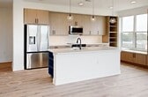 This contemporary single-floor residence offers 1,180 square feet of thoughtfully designed living space across 2 bedrooms and 2 bathrooms. Light-toned hardwood flooring flows throughout, complementing the unit's clean, modern aesthetic defined by white walls and black hardware accents.

The open-concept kitchen and living area serves as the heart of the home. The kitchen features warm wood-grain cabinetry, white quartz countertops, stainless steel appliances, a large center island with pendant lighting, open shelving, and a built-in window bench with storage — all bathed in natural light. The living area is lined with a row of large windows and a glass balcony door, providing sweeping outdoor views and direct balcony access, with a ceiling fan overhead.

The primary bedroom measures 150 square feet and includes a ceiling fan and generous windows with exterior views. It connects to a well-appointed en-suite bathroom featuring a dual-sink vanity with a bold blue cabinet, backlit mirror, glass-enclosed walk-in tile shower, and direct access to a walk-in closet with built-in shelving and hanging rods. The second bedroom also features a ceiling fan, bright windows with views, and its own closet. Additional spaces include a dedicated laundry room and two hallways connecting the layout efficiently.