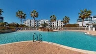 This upscale residential complex in Riverview, Florida, boasts a resort-like atmosphere centered around an expansive swimming pool. The pool area features a unique, curved design with multiple sections, including a shallow entry point and deeper swimming areas. Surrounding the pool is an extensive paved deck area with ample lounge seating, perfect for sunbathing or relaxation.

The architecture of the residential buildings is characterized by a coastal aesthetic, with white and pastel-colored exteriors, balconies, and large windows. The multi-story structures offer various apartment layouts, many with pool or landscaped views. Palm trees dot the landscape, adding to the tropical ambiance.

Community amenities include covered walkways with accessibility features, outdoor seating areas, and well-maintained landscaping. Safety features such as pool railings, emergency equipment, and clear signage are evident throughout the common areas.

The complex appears to prioritize outdoor living and social interaction, with its expansive pool area serving as a central gathering spot for residents. The overall design creates a harmonious blend of comfortable living spaces and resort-style leisure facilities, all set against the backdrop of Florida's sunny skies and lush greenery.