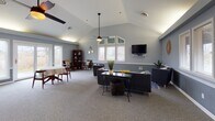 This single-floor space combines functional versatility with a cohesive, welcoming aesthetic throughout. The centerpiece is a spacious great room with soaring vaulted ceilings, soft gray carpeting, and an abundance of natural light streaming in through multiple white-trimmed windows. A wall-mounted television, ceiling fan, and comfortable seating arrangements make this area ideal for both relaxation and group gatherings. The open layout extends into an adjoining lounge area, where additional seating, a bookcase, and decorative accents contribute to the warm, lived-in ambiance. A dedicated conference room offers a polished hardwood floor, an oval wooden conference table surrounded by padded chairs, built-in shelving flanking a fireplace surround, and windows that bring in natural light — providing a professional yet inviting setting for meetings. The kitchen is well-equipped with warm wood cabinetry, stone-look countertops, stainless steel appliances including a refrigerator, range, microwave, and dishwasher, along with vinyl plank flooring that adds a clean, practical finish. Throughout the space, a consistent cool blue-gray color palette, white chair rail molding, and thoughtful décor details create a unified and polished environment suited for community, professional, or organizational use.