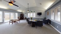 This single-floor space combines functional versatility with a cohesive, welcoming aesthetic throughout. The centerpiece is a spacious great room with soaring vaulted ceilings, soft gray carpeting, and an abundance of natural light streaming in through multiple white-trimmed windows. A wall-mounted television, ceiling fan, and comfortable seating arrangements make this area ideal for both relaxation and group gatherings. The open layout extends into an adjoining lounge area, where additional seating, a bookcase, and decorative accents contribute to the warm, lived-in ambiance. A dedicated conference room offers a polished hardwood floor, an oval wooden conference table surrounded by padded chairs, built-in shelving flanking a fireplace surround, and windows that bring in natural light — providing a professional yet inviting setting for meetings. The kitchen is well-equipped with warm wood cabinetry, stone-look countertops, stainless steel appliances including a refrigerator, range, microwave, and dishwasher, along with vinyl plank flooring that adds a clean, practical finish. Throughout the space, a consistent cool blue-gray color palette, white chair rail molding, and thoughtful décor details create a unified and polished environment suited for community, professional, or organizational use.