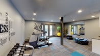 This contemporary fitness center offers a thoughtfully designed workout space spread across a single floor. The room is finished with neutral grey walls and a smooth ceiling fitted with recessed lighting, creating a clean, focused atmosphere ideal for exercise. Large glass-panel doors line the walls, inviting ample natural light and providing views to an outdoor area, enhancing the open and airy feel of the space. The flooring features a practical combination of carpet tiles with teal accent patterns and rubberized workout mats in key equipment zones. On the cardio side, the space is equipped with treadmills and elliptical trainers, while a dedicated free weights section includes a full rack of dumbbells. The functional training area is outfitted with stability balls, medicine balls, resistance equipment, and a BOX-branded training column, catering to a wide range of fitness routines. A structural column anchors the center of the room, and a large wall mirror expands the sense of space while allowing users to monitor their form. The overall ambiance is energetic yet organized, offering a well-rounded environment for both cardio and strength-based workouts.