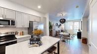 This thoughtfully designed one-bedroom, one-bathroom unit offers an efficient and functional single-floor layout with modern finishes throughout. The open-concept kitchen is a standout feature, showcasing gray shaker-style cabinetry, a white subway tile backsplash, quartz countertops, a full suite of stainless steel appliances including a French door refrigerator, electric range, built-in microwave, and dishwasher, as well as a kitchen island with bar seating — ideal for casual dining and entertaining. Light wood-style flooring runs continuously throughout the unit, lending warmth and visual cohesion. The living area is bright and inviting, benefiting from natural light through large windows and a glass door, complemented by a statement chandelier above the dining space. The bedroom is a comfortable retreat, featuring a geometric accent wall, natural light from dual windows with decorative curtains, and direct access to a dedicated closet. The bathroom is clean and well-appointed, offering a combined tub and shower, a gray vanity with a single sink, built-in wall shelving, and an arched mirror. White walls, recessed lighting, and curated wall art tie the space together for a cohesive, contemporary aesthetic.