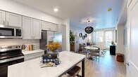 This thoughtfully designed one-bedroom, one-bathroom unit offers an efficient and functional single-floor layout with modern finishes throughout. The open-concept kitchen is a standout feature, showcasing gray shaker-style cabinetry, a white subway tile backsplash, quartz countertops, a full suite of stainless steel appliances including a French door refrigerator, electric range, built-in microwave, and dishwasher, as well as a kitchen island with bar seating — ideal for casual dining and entertaining. Light wood-style flooring runs continuously throughout the unit, lending warmth and visual cohesion. The living area is bright and inviting, benefiting from natural light through large windows and a glass door, complemented by a statement chandelier above the dining space. The bedroom is a comfortable retreat, featuring a geometric accent wall, natural light from dual windows with decorative curtains, and direct access to a dedicated closet. The bathroom is clean and well-appointed, offering a combined tub and shower, a gray vanity with a single sink, built-in wall shelving, and an arched mirror. White walls, recessed lighting, and curated wall art tie the space together for a cohesive, contemporary aesthetic.