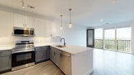 This well-appointed 1-bedroom, 1-bathroom residence offers a thoughtfully designed single-floor layout with modern finishes throughout. The open-concept kitchen and living area serves as the heart of the home, featuring two-tone cabinetry with white upper cabinets and gray lower cabinets, quartz countertops, a central island with pendant lighting, and a full suite of stainless steel appliances. Durable wood-style flooring runs throughout the main living areas, while large sliding glass doors open to a private balcony with expansive outdoor views. The spacious bedroom is carpeted for comfort and benefits from generous natural light through a large window. It includes access to a walk-in closet with built-in shelving and hanging rods. A dedicated closet off the hallway provides additional built-in shelving for extra storage. The full bathroom features a tiled shower-tub combination with a sleek sliding glass enclosure, matte black fixtures, and a vanity with ample drawer storage. A dedicated laundry room houses a stacked washer and dryer unit. The clean white walls, matte black hardware, and gray panel doors maintain a cohesive, contemporary aesthetic throughout the entire residence.
