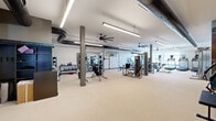 Located at 8331 S Valley Hwy in Englewood, Colorado, this well-appointed fitness center offers a generous, open floor plan designed to accommodate a comprehensive range of workout activities. The single-level space is defined by its notably high ceilings, which lend an airy, open feel throughout. Exposed ductwork and structural support columns add an industrial character, while large ceiling fans ensure consistent airflow across the workout floor.

The facility is divided into distinct functional zones. The strength training area is equipped with multiple selectorized weight machines, a squat rack loaded with plate weights, cable machines, and a dedicated free weights section featuring a full dumbbell rack. A wall of mirrors spans a large section of the space, enhancing the sense of depth and supporting proper form during exercise.

The cardio zone is outfitted with treadmills, elliptical trainers, and stationary bikes, all positioned near large windows that allow ample natural light to fill the room and offer views of the surrounding area. A separate open area provides space for functional training and stretching. Additional amenities include a small refrigerator and storage shelving. The light-toned flooring, bright linear lighting, and neutral color palette create a clean, motivating environment throughout the entire fitness center.