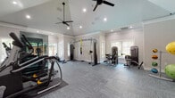 This single-story community clubhouse is thoughtfully designed to offer residents a comprehensive amenity experience. The reception area makes an immediate impression with soaring ceilings, elegant crown molding, transom windows above a grand interior archway, and light wood-look flooring that flows throughout the space. Dual wooden workstations flank the entryway, providing a polished and functional welcome area, while curated artwork adds a warm, modern touch.

Adjacent to the reception space is an open lounge area featuring large windows that invite abundant natural light, creating an airy and relaxed atmosphere. This seamlessly connects to a well-appointed kitchen with white cabinetry, a subway tile backsplash, stainless steel appliances, and a center island with a sink — ideal for community gatherings and events.

Beyond, a game room complete with a pool table offers a casual social hub. The fitness center is a standout feature, boasting a vaulted ceiling with dual ceiling fans, mirrored walls, and an extensive array of equipment including strength machines, a cable rack with TRX system, cardio machines, free weights, and stability balls — all bathed in natural light from multiple windows. The entire facility is designed to foster community, wellness, and comfort.