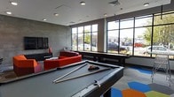 This single-floor recreational lounge is designed as a dynamic social space with a contemporary aesthetic. The room is anchored by a full-sized pool table at its center, complemented by a shuffleboard table positioned along the windowed wall — making it well-equipped for entertainment and leisure. Bold red modular seating adds a striking pop of color against the neutral gray walls and concrete-inspired finishes, while a colorful geometric area rug introduces additional visual energy to the space. Large, industrial-style floor-to-ceiling windows line one side of the room, flooding the interior with abundant natural light and offering views of the surrounding streetscape. Recessed ceiling lighting supplements the natural light, ensuring the space remains well-lit throughout the day. A wall-mounted flat-screen television is positioned near the lounge seating area, adding to the room's versatility as a gathering space. The overall ambiance is modern and lively, blending industrial design elements with vibrant accent pieces to create an inviting, activity-focused environment suited for socializing and recreation.