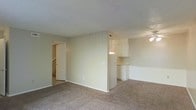 This two-story residence offers a straightforward and functional layout designed for comfortable everyday living. The main level features a spacious living area finished with wall-to-wall carpeting in a neutral tone, creating a warm and understated atmosphere throughout. The open floor plan allows the living space to flow naturally toward the kitchen, which is partially separated by a dividing wall that adds a sense of definition without fully closing off the area. The kitchen is outfitted with white cabinetry and a white refrigerator, contributing to a clean, bright aesthetic. A ceiling fan is visible overhead, providing practical ventilation and lighting to the adjoining space. A doorway from the main living area leads to a staircase accessing the second floor, suggesting additional living or sleeping quarters above. HVAC vents are integrated into the ceiling, ensuring climate control throughout. The overall palette of the space is light and neutral, with beige walls and white trim that lend a cohesive and airy feel. The layout is efficient and well-suited for a variety of living arrangements, with clear sightlines between the primary living zone and kitchen area making the home feel open and connected across both levels.