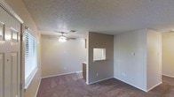 This single-floor residence offers a straightforward and functional layout designed for comfortable everyday living. The main living area is finished with plush carpet flooring that adds warmth underfoot, while a ceiling fan with a built-in light fixture provides both air circulation and ambient lighting. Neutral-toned walls in soft beige and taupe create a calm, cohesive atmosphere throughout the space.

A defining architectural detail is the partial dividing wall featuring a pass-through window, which visually connects the living area to the kitchen beyond. This opening allows for natural sightlines between the two spaces, enhancing the sense of openness while maintaining a defined separation between areas. The kitchen can be glimpsed through the pass-through, where cabinetry is visible, suggesting a compact and efficiently arranged cooking space.

The textured popcorn ceiling adds a classic touch and runs consistently throughout, contributing to the overall character of the interior. Ample wall outlets are distributed across the living area, offering practical convenience. The neutral color palette and carpeted floors lend the space a quiet, understated quality that provides a versatile backdrop for a variety of interior styles and personal touches.