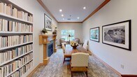 Located at 1301 W Whitestone Blvd in Cedar Park, Texas, this single-floor space combines a warm, sophisticated aesthetic with functional design. The main lounge area serves as the centerpiece, anchored by a classic wood-mantled fireplace with a blue-tiled surround. Built-in bookshelves flank the fireplace, providing ample storage and a scholarly ambiance. A rustic solid-wood dining or conference table sits atop a patterned area rug, surrounded by upholstered chairs, making it ideal for gatherings or collaborative meetings. A pair of cushioned armchairs positioned near a large window creates an inviting reading nook bathed in natural light, with views of the lush, tree-lined outdoor surroundings. Recessed lighting and warm wood crown molding throughout lend a polished, cohesive finish to the interior. Framed black-and-white landscape photographs adorn the walls, adding an artistic and curated touch. An adjoining area features a dedicated workstation with ergonomic seating and a computer setup, accessible through a glass-paned French door that allows light to flow between spaces. Neutral wall tones, warm wood trim, and carpeted flooring tie the entire space together, creating a professional yet welcoming environment well-suited for office, community, or business use.