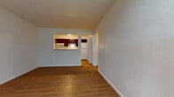 This single-floor unit offers a functional and well-organized layout across several distinct spaces. The living area is generously sized, finished with warm hardwood-style flooring throughout, and features a wall-mounted HVAC unit alongside a large window that brings in natural light and views of surrounding greenery. A pass-through opening connects the living area to the kitchen, creating an open and communicative feel between the two spaces. The kitchen is equipped with rich, dark-stained cabinetry spanning both upper and lower sections, a white electric range, a full-size refrigerator, and a single-basin sink — all set atop matching hardwood-style flooring for visual continuity. A hallway leads from the main living space to the additional rooms. The bedroom features a colorful patchwork carpet, a baseboard heater, and a window with blinds overlooking lush outdoor foliage, providing both warmth and a connection to the outdoors. The bathroom is clean and efficiently designed, offering a full tub-shower combination with built-in shelving, a white vanity with ample cabinet storage, brushed nickel fixtures, and hardwood-style flooring. The overall aesthetic is practical and neutral, with light-toned walls creating a bright, move-in ready environment throughout.