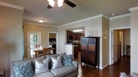 This single-floor residence offers a thoughtfully designed open-concept layout that seamlessly connects the main living spaces. The living area is anchored by a plush sofa accented with decorative pillows and features rich hardwood flooring that flows throughout the home. A ceiling fan overhead adds both style and comfort to the space. Crown molding along the upper walls lends a refined, classic touch to the overall aesthetic.

Adjacent to the living area, the kitchen is defined by warm, dark wood cabinetry and a convenient pass-through counter that maintains a sense of openness between the spaces. The kitchen is well-equipped and efficiently laid out, making it both practical and visually appealing.

A dedicated dining nook is situated just off the living area, bathed in natural light streaming through French doors that open to the outdoors, providing a bright and inviting atmosphere for meals. The connection between interior and exterior gives the space a welcoming, airy feel.

Throughout the home, neutral wall tones complement the warm wood elements, creating a cohesive and comfortable ambiance. The single-floor design ensures ease of movement and accessibility, making this a practical and attractive living space suitable for a variety of lifestyles.