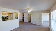 This single-floor residence offers a straightforward and functional layout designed for comfortable everyday living. The main living area is generously sized, featuring wall-to-wall carpeting and a ceiling fan with integrated lighting that anchors the room. A textured ceiling adds a classic touch throughout the space.

A built-in desk nook along one wall provides a dedicated workspace, making the living area both versatile and practical. The kitchen connects to the living space via a pass-through counter with overhead cabinetry visible on the other side, allowing for an open and interactive feel between the two areas. A small overhead light illuminates the kitchen pass-through, adding warmth to the transition zone.

Multiple doorways lead from the main living area, suggesting access to bedrooms, bathrooms, and additional storage or utility spaces. The hallways are carpeted and well-lit, maintaining visual continuity throughout the home. White walls and neutral tones create a clean, bright atmosphere that makes the space feel open and easy to personalize. Overall, this residence combines practical design elements with a comfortable, livable ambiance suited for a variety of lifestyles.