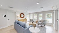 This 410-square-foot single-floor space is a well-appointed community lounge comprising a living room and a dining area. The interior is finished with light-toned luxury vinyl plank flooring throughout, neutral beige walls, and recessed lighting, creating a clean, welcoming atmosphere.

The living room is the centerpiece of the space, featuring multiple large windows with white horizontal blinds that allow abundant natural light to fill the room. A glass-paneled exterior door provides direct access to an outdoor area, further enhancing the bright, airy feel. Seating includes a light blue sofa and patterned accent chairs arranged around a round natural wood coffee table atop a geometric area rug. A wooden sideboard console along one wall is topped with a decorative starburst mirror, adding a distinctive focal point.

Adjoining the living room is a compact wet bar or kitchenette area with white upper cabinetry, a sink, and bar-height seating — ideal for casual gatherings. The dining area is a cozy, defined nook featuring a round white dining table surrounded by upholstered chairs, a window with garden views, and a vibrant abstract wall print that adds a pop of color. Together, the rooms offer a cohesive, coastal-inspired aesthetic suited for relaxed communal use.