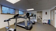 This fitness center in Delafield, Wisconsin offers a well-equipped, single-floor exercise facility designed to support a range of workout routines. The space is organized into distinct zones, each serving a specific training purpose. The main cardio area features multiple treadmills, including LifeSpan models, arranged in a row and complemented by large wall-mounted mirrors that visually expand the space and allow users to monitor their form. A drop ceiling with recessed fluorescent lighting provides consistent, even illumination throughout, while small windows along one wall bring in a measure of natural light. A secondary area houses stationary bikes, including both an upright spin bike and a recumbent cycle, alongside a weight bench and additional resistance equipment. This zone also features wall-mounted mirrors and a wall-unit air conditioner for climate control. A dedicated strength training room contains a full barbell rack with weight plates, a stability ball, and a workout bench, offering a more enclosed environment for focused lifting sessions. A wall-mounted television is available in the main workout area, and a coat rack near the entrance provides convenience for users. The entry point features a glass-paneled door, a welcome mat, a body weight scale, and a fire extinguisher, reflecting a practical and safety-conscious design. The carpeted flooring throughout adds comfort underfoot during workouts.