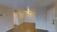 This single-floor space presents a clean, open layout with several notable architectural details. The living area is distinguished by warm-toned parquet flooring arranged in a classic geometric pattern, adding visual texture and character underfoot. The ceiling features a textured finish and is equipped with a ceiling fan fitted with a light fixture, providing both ventilation and ambient lighting throughout the space. HVAC ventilation is integrated into the ceiling, ensuring efficient climate control. The walls are finished in a neutral, light tone, creating a bright and versatile backdrop. White baseboards run along the perimeter, adding a crisp, defined contrast to the flooring. A doorway visible in the corner leads to an adjoining room or closet, suggesting a functional and organized layout. A partial wall or column detail is visible on the right side of the space, hinting at a defined transition between areas. Overall, the space offers a straightforward, well-maintained interior with classic finishes that provide a neutral canvas suitable for a variety of uses and furnishing styles.
