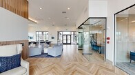 This thoughtfully designed single-story space offers a generous, open-plan layout that seamlessly integrates multiple functional zones within its expansive footprint. The interior is defined by light wood herringbone-patterned flooring that transitions into wide-plank hardwood throughout the main living areas, lending a cohesive and refined aesthetic.

The living area features soaring ceilings with vertical wood slat wall paneling as a prominent design element, anchoring a curated seating arrangement of neutral sofas and accent chairs. Tall, narrow windows along the perimeter flood the space with natural light and provide outdoor views, while a large wall-mounted display and contemporary chandelier add to the polished atmosphere.

The kitchenette area is appointed with a white quartz waterfall island, bar seating with tufted blue chairs, warm wood cabinetry, and a built-in coffee station beneath sculptural pendant lighting.

Flanking the main open area are fully enclosed glass-walled rooms with black metal framing, housing a conference-style room with a wood table and blue chairs, and a private office with a traditional desk setup—offering transparency while maintaining acoustic separation. The overall ambiance is modern, airy, and professionally styled.