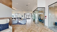 This thoughtfully designed single-story space offers a generous, open-plan layout that seamlessly integrates multiple functional zones within its expansive footprint. The interior is defined by light wood herringbone-patterned flooring that transitions into wide-plank hardwood throughout the main living areas, lending a cohesive and refined aesthetic.

The living area features soaring ceilings with vertical wood slat wall paneling as a prominent design element, anchoring a curated seating arrangement of neutral sofas and accent chairs. Tall, narrow windows along the perimeter flood the space with natural light and provide outdoor views, while a large wall-mounted display and contemporary chandelier add to the polished atmosphere.

The kitchenette area is appointed with a white quartz waterfall island, bar seating with tufted blue chairs, warm wood cabinetry, and a built-in coffee station beneath sculptural pendant lighting.

Flanking the main open area are fully enclosed glass-walled rooms with black metal framing, housing a conference-style room with a wood table and blue chairs, and a private office with a traditional desk setup—offering transparency while maintaining acoustic separation. The overall ambiance is modern, airy, and professionally styled.