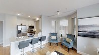 This single-floor space showcases a thoughtfully designed open-concept layout that seamlessly connects the kitchen and living area. The kitchen is outfitted with sleek white cabinetry, stainless steel appliances including a refrigerator, microwave, and range, and a clean white countertop peninsula with bar seating for casual dining. The peninsula, lined with contemporary gray upholstered barstools on chrome bases, serves as a natural divider between the kitchen and living space.

The living area is bright and airy, bathed in natural light streaming through multiple windows fitted with white horizontal blinds. Two plush blue-gray wingback chairs with dark wood legs anchor a cozy seating nook, complemented by a small round side table adorned with a floral arrangement. The recessed lighting throughout enhances the overall brightness of the space.

Light-toned hardwood flooring runs throughout, adding warmth and continuity to the crisp, neutral color palette of white and soft gray. The combination of modern finishes, natural light, and an efficient open layout gives this space a clean, contemporary ambiance well-suited for everyday living.