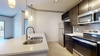 This single-floor residence in Louisville, KY, showcases a thoughtfully designed interior with contemporary finishes throughout. The kitchen serves as a central highlight, featuring rich dark-stained cabinetry that extends to the ceiling, complete with a built-in wine rack. Stainless steel appliances—including a refrigerator, electric range, and over-the-range microwave—are complemented by a mosaic tile backsplash and light quartz countertops. A large peninsula with a double undermount sink provides additional workspace and casual seating, illuminated by elegant pendant lighting suspended from a track-lit ceiling. Light-toned wood-look flooring flows throughout the kitchen area, adding warmth to the space.

The bedroom areas feature plush carpeting and neutral white walls, creating a calm and versatile environment. Panel-style doors in a soft gray tone add a modern architectural detail, while windows fitted with blinds allow for controlled natural light.

The bathroom is well-equipped with a dark wood vanity, white quartz countertop, and a large frameless mirror above, offering ample storage through multiple drawers and cabinet space.

A private balcony with a black metal railing extends the living space outdoors, offering open views of the surrounding building exteriors under open skies.