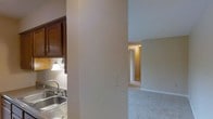 This single-story space is characterized by a straightforward, functional layout that connects its key areas through an open corridor design. The kitchen, visible to one side, features dark wood cabinetry that provides a warm contrast to the neutral wall tones. A stainless steel sink sits beneath a wall-mounted light fixture, which casts a warm glow over the workspace, enhancing the functional nature of the area. The cabinetry offers ample storage and contributes a rich, traditional aesthetic to the kitchen zone.

A structural column serves as a subtle divider between the kitchen and the adjoining living area, helping to define each space while maintaining an open feel. The living area is finished with wall-to-wall carpeting in a neutral tone, lending a soft, comfortable atmosphere underfoot. The walls throughout are painted in light, muted shades that help reflect available light and create a cohesive, calm ambiance.

A hallway visible in the background leads to additional rooms, hinting at a thoughtfully connected floor plan. White door trim and baseboards provide clean, classic detailing throughout, complementing the overall understated and practical character of the space.