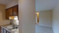 This single-story space is characterized by a straightforward, functional layout that connects its key areas through an open corridor design. The kitchen, visible to one side, features dark wood cabinetry that provides a warm contrast to the neutral wall tones. A stainless steel sink sits beneath a wall-mounted light fixture, which casts a warm glow over the workspace, enhancing the functional nature of the area. The cabinetry offers ample storage and contributes a rich, traditional aesthetic to the kitchen zone.

A structural column serves as a subtle divider between the kitchen and the adjoining living area, helping to define each space while maintaining an open feel. The living area is finished with wall-to-wall carpeting in a neutral tone, lending a soft, comfortable atmosphere underfoot. The walls throughout are painted in light, muted shades that help reflect available light and create a cohesive, calm ambiance.

A hallway visible in the background leads to additional rooms, hinting at a thoughtfully connected floor plan. White door trim and baseboards provide clean, classic detailing throughout, complementing the overall understated and practical character of the space.