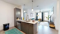 This thoughtfully designed residence offers five bedrooms and four bathrooms across a single floor, making it an excellent option for shared living. The open-concept layout connects the kitchen and living room seamlessly, creating a bright, communal hub at the heart of the home. The kitchen is outfitted with stainless steel appliances, including a refrigerator, range, and built-in microwave, complemented by a large peninsula with a deep single-basin sink, pendant lighting overhead, white upper cabinetry, and dark lower cabinets with light countertops — ideal for both cooking and casual dining. The living area features light wood-look flooring, a ceiling fan, and large windows with a glass door opening to a private balcony, allowing ample natural light to fill the space. The bedrooms are well-proportioned, each with closet storage and wood-look flooring; the primary bedroom includes its own dedicated closet and a window with exterior views. The bathrooms are clean and modern, featuring subway-tiled tub-shower combos, wood-finish vanities, and white quartz countertops. Throughout the residence, the neutral color palette and contemporary finishes create a cohesive, functional living environment suited to multiple residents.