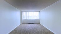 This single-floor space is designed with simplicity and functionality in mind, offering a clean, open layout that maximizes usable living area. The main living space is finished with wall-to-wall neutral-toned carpeting, providing a soft and comfortable underfoot feel throughout. Crisp white walls and a smooth white ceiling contribute to a bright, minimalist aesthetic that lends itself well to a variety of interior styles.

A standout feature of the living area is the expansive picture window spanning much of the exterior wall. Dressed with sheer vertical blinds, this large window allows an abundance of natural light to flood the interior while offering views of the surrounding greenery visible through the glass. A baseboard heating unit positioned beneath the window ensures comfortable climate control during cooler months.

The overall ambiance of the space is calm and neutral, providing a versatile canvas for personalization. The absence of interior clutter highlights the generous proportions of the room and the clean architectural lines of the space. White baseboards frame the perimeter of the room, adding a subtle finishing touch that ties together the light, cohesive color palette throughout.