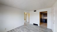 This single-floor space in Fairway offers a straightforward, functional layout designed for comfortable everyday living. The main living area features light gray wood-look flooring that extends throughout, providing a clean and cohesive aesthetic. Bright white walls reflect natural and artificial light, contributing to an airy, open feel throughout the space.

A wide doorway opening from the main living area leads directly to the kitchen, which is finished with dark wood cabinetry and tile flooring, creating a subtle contrast with the lighter tones of the living space. The kitchen includes a sink and appears to offer ample lower cabinet storage. An additional doorway from the main area connects to a hallway or secondary room, suggesting a practical flow between spaces.

Ceiling-mounted light fixtures provide consistent overhead lighting, and a centrally located HVAC vent ensures climate control throughout. The neutral color palette and uncomplicated layout make this space adaptable to a variety of furniture arrangements and interior styles.