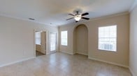 This single-floor space offers a clean and inviting atmosphere defined by thoughtful architectural details and a warm, neutral color palette. The main living area immediately draws the eye with its elegant arched wall niche, a charming decorative feature that adds character and visual depth to the room. Crown molding lines the ceiling, lending a polished, classic finish to the space. A ceiling fan with a built-in light fixture is centrally positioned, providing both style and practical airflow. The room is finished with large-format ceramic tile flooring in a soft, light tone that extends throughout, creating a seamless and easy-to-maintain surface underfoot. Multiple windows fitted with horizontal blinds allow generous amounts of natural light to fill the space, enhancing its bright and open feel. A glass-paneled exterior door further contributes to the light-filled ambiance while offering direct access to the outdoors. An adjoining room is visible through an open doorway, suggesting a well-connected and functional floor plan. The overall aesthetic is understated and versatile, making the space well-suited for a variety of uses while maintaining a comfortable and welcoming environment.