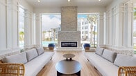 Located at 1799 North Williamson Boulevard in Daytona Beach, FL, this single-level clubhouse lounge offers a refined coastal retreat designed for relaxation and social gathering. The open-concept living room is anchored by a striking floor-to-ceiling stacked stone fireplace, which serves as the architectural centerpiece of the space. Light wood-look flooring runs throughout, complementing the bright, airy palette of crisp whites and soft coastal blues that define the interior aesthetic.

Oversized columns and wide open-air passages seamlessly connect the indoor lounge to a covered outdoor terrace, dissolving the boundary between interior comfort and the vibrant poolside environment beyond. Large glass panel walls and folding door systems flood the space with natural light and provide direct visual access to the resort-style pool, lined with lounge chairs and swaying palm trees.

The living area is furnished with a curated mix of plush sofas, woven rattan chairs, and low-profile coffee tables, striking a balance between casual comfort and upscale design. Ceiling fans ensure year-round comfort in the covered outdoor sections. A separate interior room, visible through full-height glass bi-fold doors with transom windows above, offers an additional enclosed gathering space. The overall ambiance is that of a sophisticated, tropical-inspired amenity lounge befitting the Florida lifestyle.