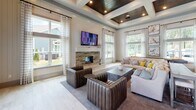 Located at 50 Cheryl Lane in Summerville, SC, this single-story community clubhouse is designed with a refined yet welcoming aesthetic that blends modern farmhouse and coastal influences throughout. The grand great room serves as the heart of the space, boasting soaring coffered ceilings with rich wood inlays, recessed lighting, and an eye-catching stone fireplace flanked by a wall-mounted TV. Expansive windows dressed in crisp striped drapery flood the room with natural light and offer views of the surrounding greenery, creating an airy, open ambiance. The fully equipped community kitchen features slate-blue shaker cabinetry, white quartz countertops, a mosaic tile backsplash, a large center island with seating, stainless steel appliances, and dramatic industrial-style pendant lighting. A dedicated conference room provides a functional gathering space with a rustic wood table, classic Windsor-style chairs, shiplap-accented walls, and a wall-mounted display screen. The entry foyer is appointed with large-format tile flooring and framed nature-inspired artwork, with glass double doors that open directly to the outdoor pool area. The cohesive design palette, featuring neutral tones, warm wood accents, and curated artwork, lends the entire space a polished yet approachable character.
