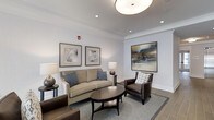 This single-floor space presents a thoughtfully designed lounge area that balances comfort and elegance. The room features a neutral color palette dominated by soft whites and warm grays, creating a calm and welcoming atmosphere. Crown molding lines the ceiling perimeter, adding a refined architectural detail that enhances the overall sense of polish.

The seating arrangement centers around a curved beige sofa accented with blue throw pillows, paired with a dark brown leather armchair featuring a geometric-patterned accent pillow. A sleek, oval dark wood coffee table anchors the seating group, complemented by a coordinating area rug that defines the space. Table lamps on either side of the arrangement provide warm ambient lighting, supplemented by recessed ceiling lights and a prominent flush-mount drum light fixture overhead.

Wall decor includes a pair of framed abstract prints and a larger statement canvas in earth and blue tones, adding visual interest without overwhelming the serene environment. The flooring transitions from the area rug to what appears to be a light hardwood or tile border, further enhancing the room's structured, upscale feel.
