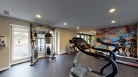 This single-level fitness center in San Antonio, Texas offers a thoughtfully designed workout environment suited for a range of exercise routines. The space is divided into two distinct zones: a primary exercise area with rubber flooring that houses the majority of the equipment, and a secondary section with warm hardwood flooring ideal for stretching, yoga, or bodyweight training—complete with a yoga mat and resistance accessories.

The main workout area is equipped with multiple treadmills, a recumbent bike, an upright stationary bike, and a multi-function cable resistance machine, providing options for both cardiovascular and strength training. A wall-mounted flat-screen television is positioned for easy viewing during workouts. Recessed lighting throughout keeps the space well-lit, while large windows along the walls invite natural light and outdoor views, contributing to an open, energizing atmosphere.

A bold, colorful geometric mural serves as a striking focal point on one accent wall, adding visual energy to the space. Neutral-toned walls and a textured ceiling complete the clean, functional aesthetic. The layout is practical and spacious, allowing users to move freely between equipment. The combination of dedicated flooring zones, diverse equipment, and vibrant design elements makes this fitness center a well-rounded facility.