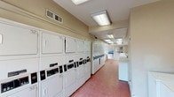 This laundry center is a single-floor facility designed to accommodate a high volume of laundry needs efficiently. The space is organized as a long, open room with machines lining both sides, creating a clear central aisle that allows for easy movement throughout. On one side, a row of stacked commercial dryers runs the length of the room, maximizing vertical space and overall drying capacity. Opposite, top-loading and front-loading washers are arranged in a continuous line, each equipped with electronic control panels for straightforward operation. Informational posted signage, including washing instructions and usage tips, is visible along the walls to assist users. The floor is finished in durable tile with a warm terracotta tone, suited to the practical demands of the space. Overhead, recessed fluorescent lighting provides consistent, bright illumination throughout the room. A large window and glass door at one end allow natural light to enter, contributing to an open and functional atmosphere. Ventilation is supported by ceiling vents distributed along the room. An electrical panel is mounted on the wall near the entrance, and open counter space is available for folding and sorting laundry. The layout and equipment volume reflect a facility built to serve a large community efficiently.