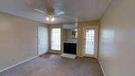 This single-floor, one-bedroom, one-bathroom unit offers a functional and straightforward layout. The main living area serves as the focal point of the space, centered around a fireplace with a raised hearth, adding a cozy element to the room. Carpet flooring extends throughout the living area, providing a warm underfoot feel, while a section of hard flooring is visible near the entryway. Two windows flank the fireplace, allowing natural light to filter into the space and creating a bright, open atmosphere. A ceiling fan with a built-in light fixture is mounted overhead on a textured popcorn ceiling, offering both air circulation and additional lighting. The room features two exterior-facing doors, enhancing accessibility and adding to the flow of natural light. A mirrored wall panel is positioned above the fireplace, visually expanding the space and reflecting light throughout the room. Neutral-toned walls complement the carpeting, contributing to a clean and understated aesthetic throughout the unit.