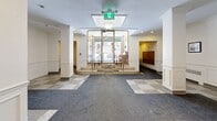 Located at 10 Rosemount in the prestigious Westmount neighbourhood of Quebec, this building lobby offers a welcoming and well-appointed entry experience. The space is defined by its classic architectural character, featuring crisp white wainscoting, decorative pilasters, and crown moulding that lend a timeless, refined aesthetic throughout.

The main entry vestibule is a standout feature, framed by glass panels and brass railings with elegant stone steps leading inward, allowing natural light to filter into the heart of the lobby. A stainless steel elevator is centrally positioned and easily accessible, flanked by the decorative pilasters that add visual structure to the space.

The flooring presents an attractive combination of a terrazzo tile checkerboard pattern near the entry and darker carpet in the main circulation areas, creating a clear and functional flow. Recessed lighting and flush-mounted fixtures illuminate the space evenly, complementing the warm, neutral wall tones.

Additional details include a large wood-framed interior window that connects to an adjacent room, framed artwork displayed along the walls, and ornate gold-framed mirrors that enhance the sense of space. The overall ambiance is polished and inviting, reflecting the distinguished character expected of a Westmount address.