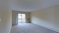 This single-floor unit offers a straightforward and functional layout centered around a spacious living area. The room is finished with neutral beige walls and wall-to-wall carpeting in a light grey tone, creating a clean and understated aesthetic throughout.

A sliding glass door serves as the focal point of the living space, filling the room with natural light and providing direct access to a private balcony. Views of surrounding trees and an adjacent building are visible through the glass, offering a glimpse of the outdoor environment.

Climate control is handled through a combination of a wall-mounted air conditioning unit positioned beneath the sliding door and baseboard heating running along the perimeter of the room, ensuring comfort year-round. The ceiling is smooth and painted white, contributing to the bright, open feel of the interior.

The overall ambiance is simple and move-in ready, with a neutral palette that lends itself well to a variety of furnishing styles. The space is practical and well-lit, making it a functional living environment suitable for everyday use.