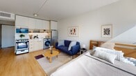 Located at 161 W 600 S in Salt Lake City, Utah, this single-floor studio is designed with a clean, modern aesthetic that maximizes its open layout. Light wood flooring runs throughout the entire space, lending warmth and visual continuity from the entry corridor to the living and sleeping areas.

The kitchen is positioned along one wall and features white cabinetry with under-cabinet lighting, a stainless steel refrigerator, an electric range, a black-finish faucet, and ample counter space. An in-unit washer is integrated seamlessly alongside the kitchen appliances, adding practical convenience.

The living area accommodates a compact sofa, a round wood coffee table, and an area rug, creating a defined lounge zone within the open floor plan. A wall-mounted flat-screen TV and a multi-drawer dresser are positioned opposite the sleeping area, which includes a wood-headboard bed framing and additional storage.

A large floor-to-ceiling window at the far end of the space draws in generous natural light and provides views of the surrounding Salt Lake City cityscape, enhancing the bright, airy ambiance. An arched mirror detail near the entry adds architectural character. The overall design is minimal, functional, and cohesive throughout.