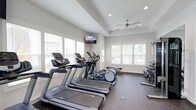 Located at 2440 Salinger Ct in Wilmington, NC, this fitness center offers a well-appointed, single-floor exercise facility designed to support a comprehensive workout routine. The space is bathed in natural light streaming through multiple large windows fitted with white blinds, creating a bright and energizing atmosphere. A tray ceiling with recessed lighting and a ceiling fan ensures the room remains comfortable and well-lit during any workout session. The cardio area is generously equipped with multiple treadmills and elliptical machines arranged in orderly rows, providing ample space for users to exercise without feeling crowded. A wall-mounted flat-screen television adds an entertainment element, making longer cardio sessions more engaging. On the opposite side of the room, a dedicated free weights section features a rack of dumbbells in varying sizes alongside additional strength training equipment, catering to a range of fitness levels. The durable, dark-toned flooring throughout the space is well-suited for high-traffic use and complements the clean, neutral color palette of the walls. Overall, the fitness center strikes a balance between functionality and comfort, offering residents a welcoming and fully equipped environment to pursue their health and wellness goals.