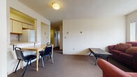 This single-floor unit is designed with practicality in mind, offering a straightforward layout that accommodates shared living comfortably. The combined living and dining area serves as the central hub of the unit, where a dining table with seating for several is positioned adjacent to a kitchenette. The kitchenette includes upper cabinetry, a countertop with an integrated sink, and a full-size refrigerator — providing essential cooking and storage functionality. The living area features carpet flooring throughout, a textured ceiling with flush-mount lighting, and a large window that fills the space with natural light and offers views of the surrounding neighborhood. Two sofas and a coffee table furnish the sitting area, creating a casual gathering space.

Each bedroom is carpeted and similarly appointed, featuring large windows that bring in ample natural light and frame exterior views. The rooms accommodate a bed, dresser, and desk workspace. Baseboard heating units are present in each bedroom. The bathroom is clean and efficient, featuring a single vanity with storage, a toilet, and a tub-shower combination with a sliding glass door and mirrored medicine cabinet above.